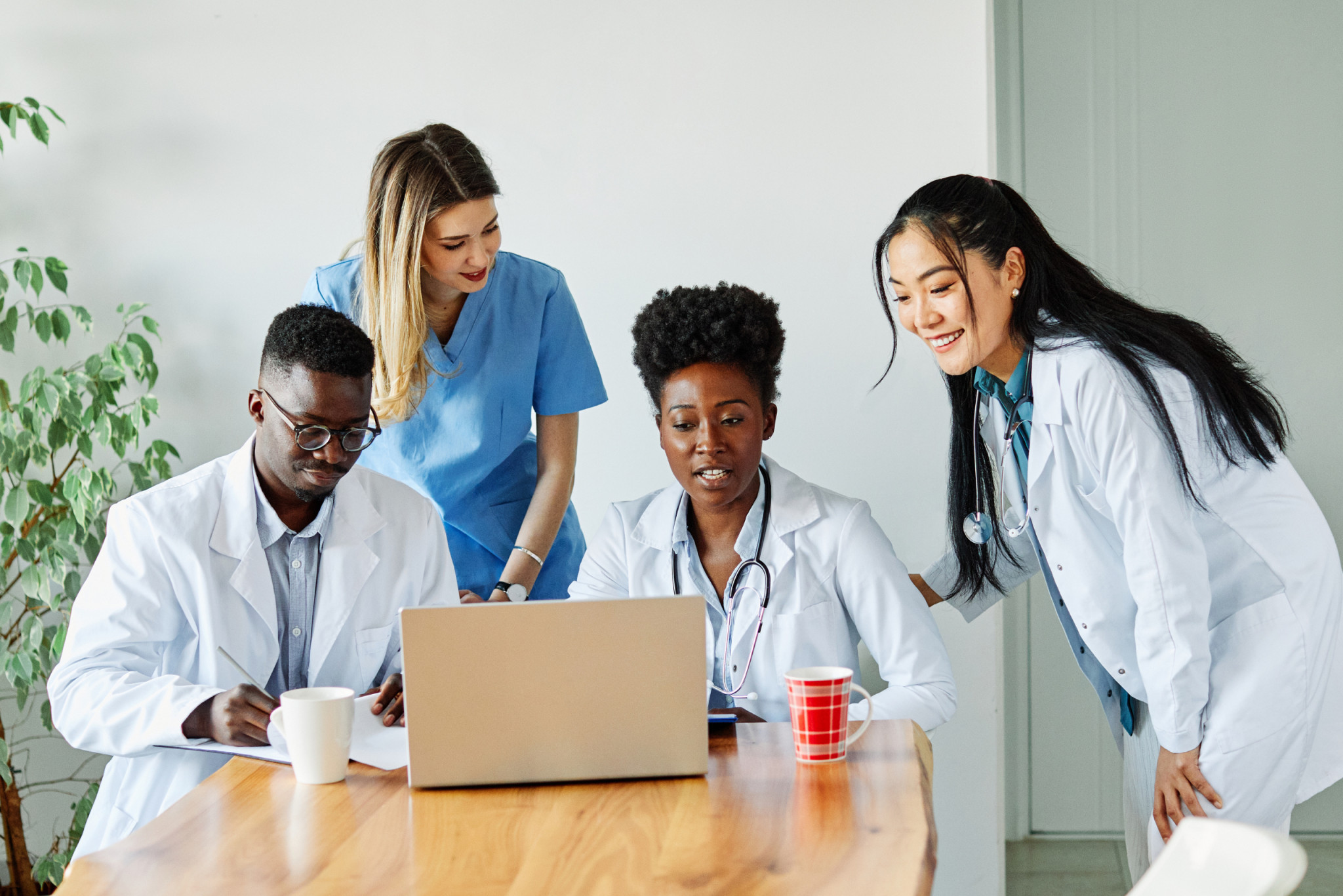 Four clinicians in white coats and scrubs gathered around a laptop at a conference table