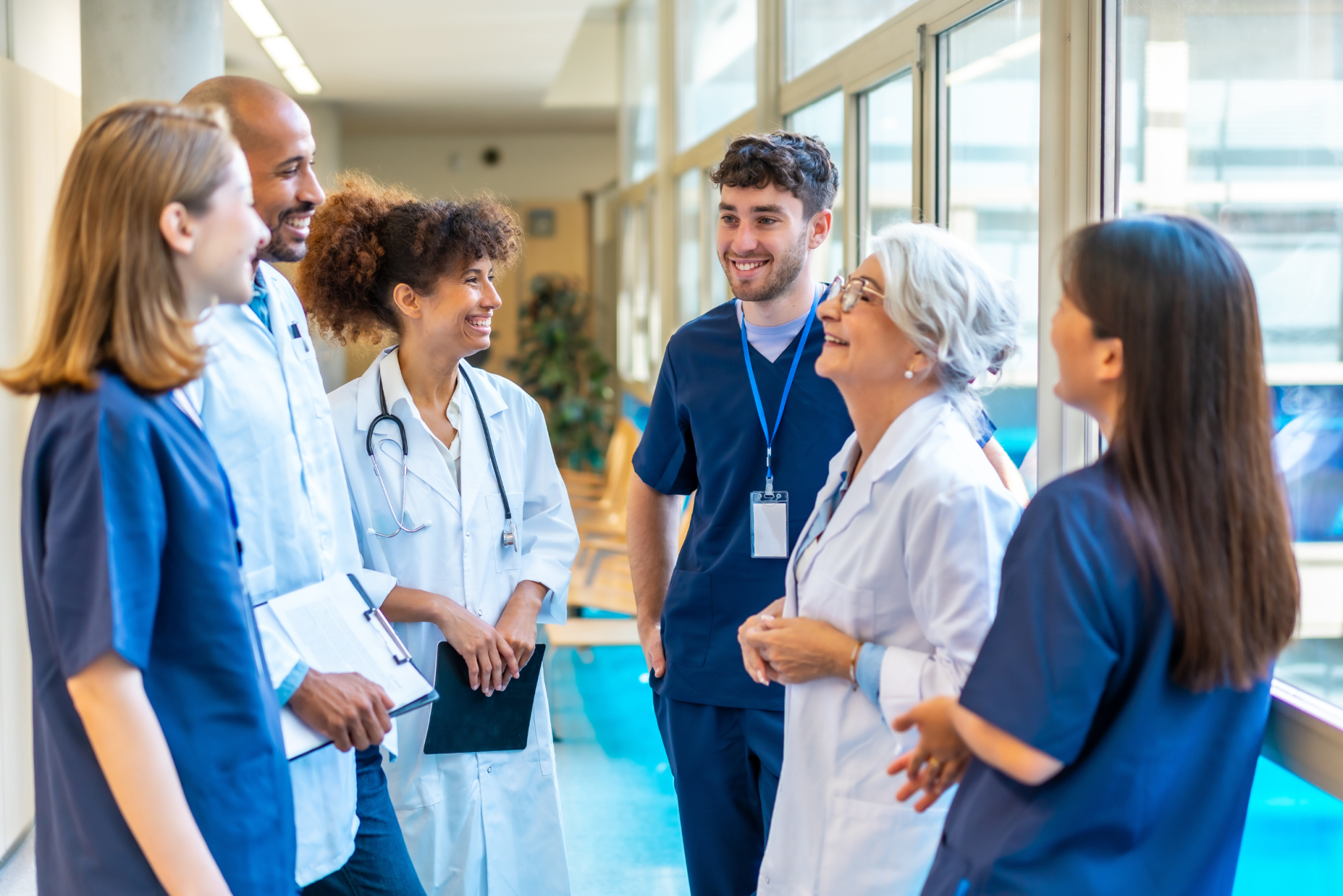Group of smiling nurses and doctors in scrubs and white coats talking together in a bright hospital hallway