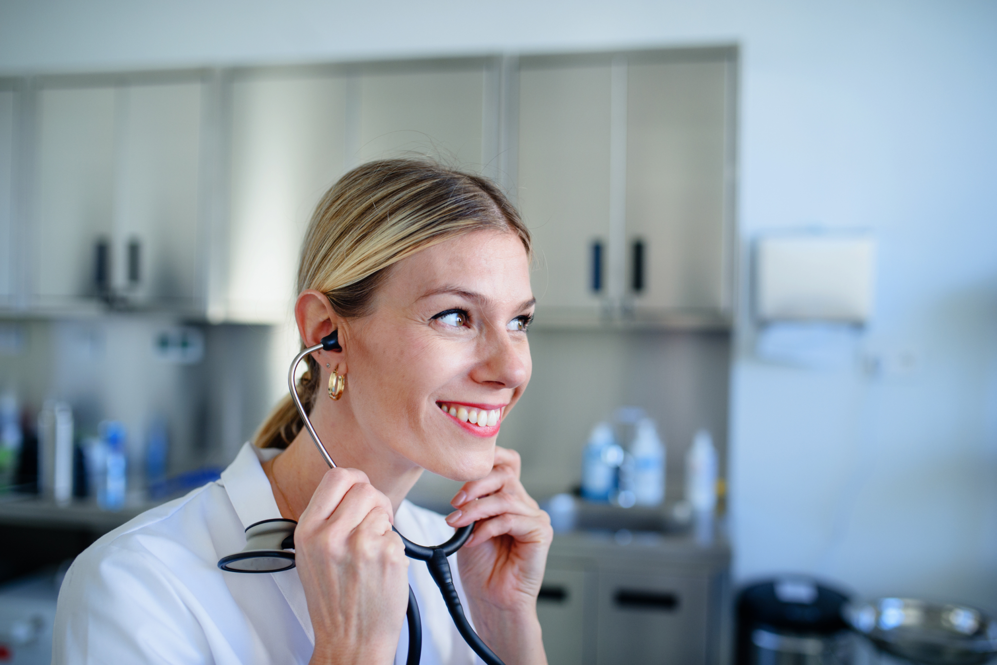 Smiling clinician in a white coat putting on a stethoscope in a clinical workroom